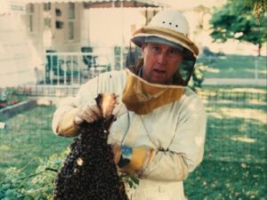 Beekeeper in protective gear holding a cluster of bees, showcasing beekeeping practices in a garden setting.