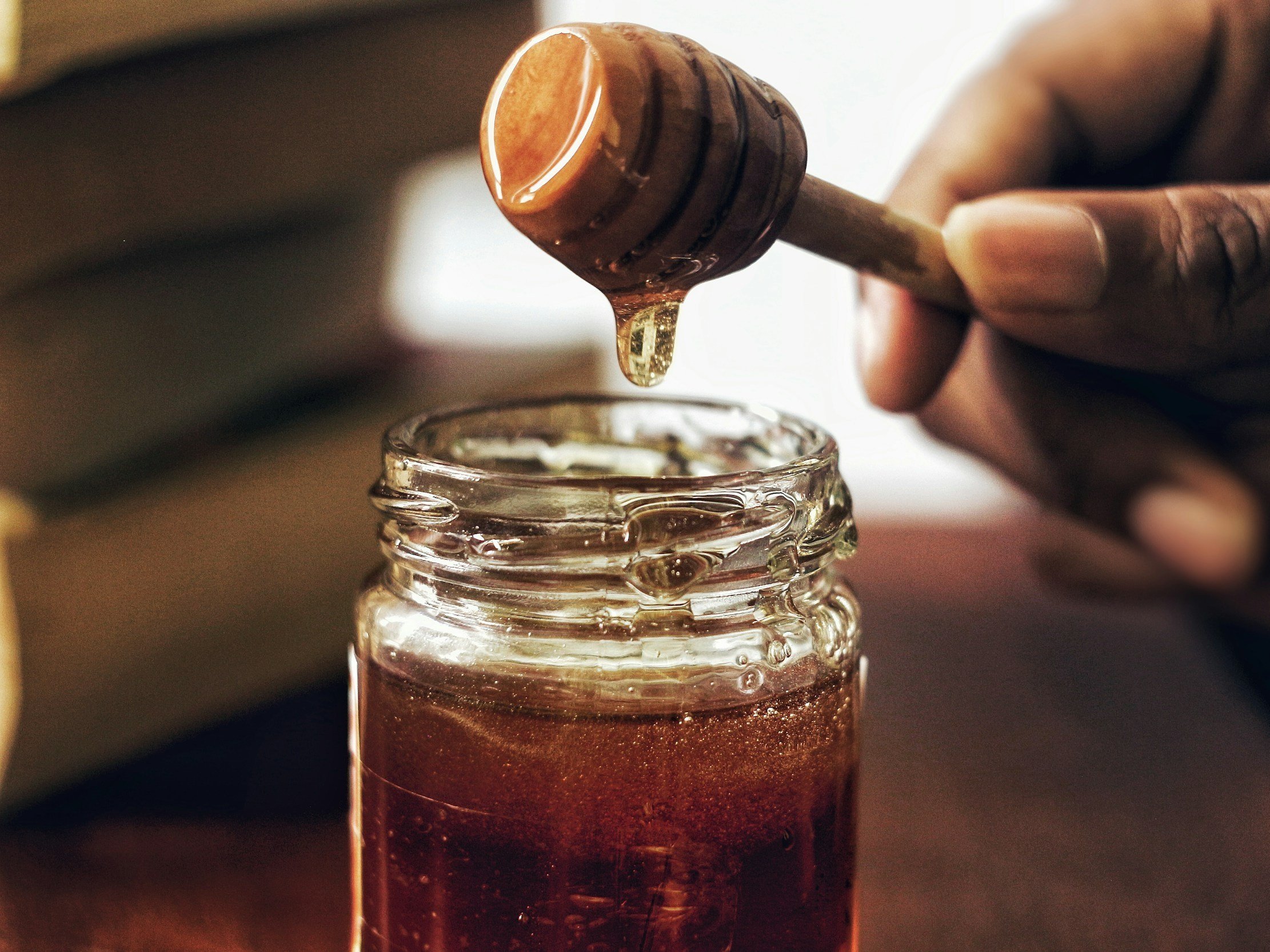 Close-up of a hand holding a honey dipper above a glass jar filled with golden honey, with books blurred in the background, highlighting the natural sweetness and texture of honey.